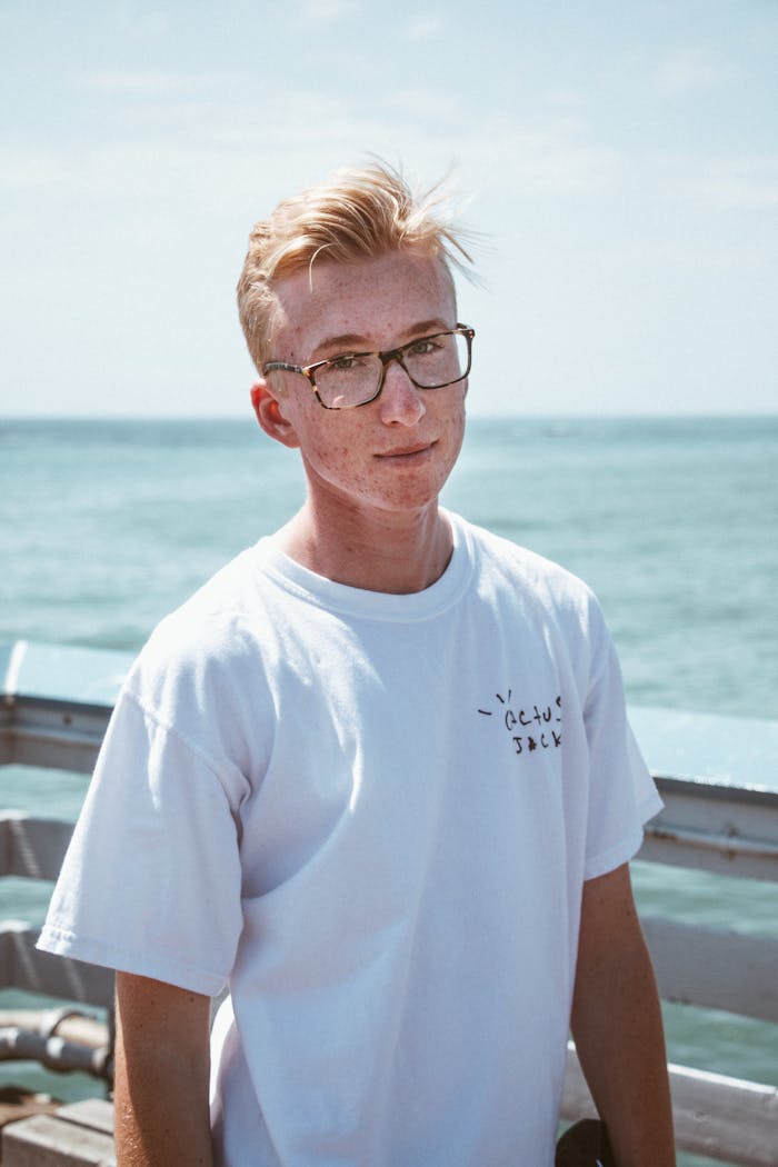 about-us Young man in glasses wearing a white shirt, standing by the sea on a sunny day.